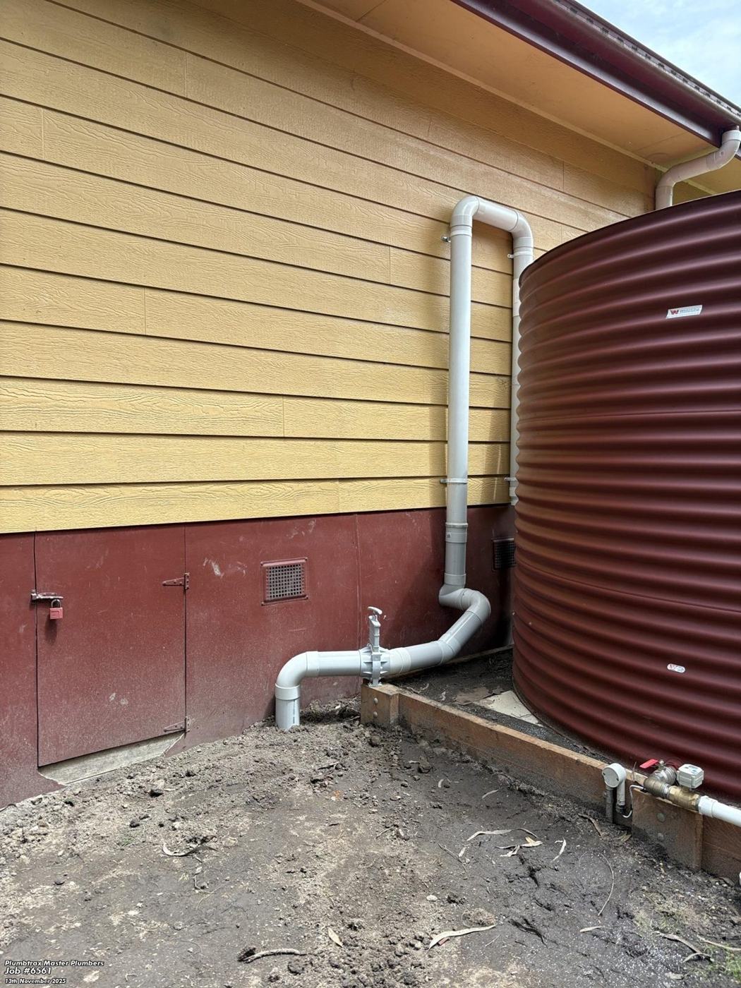 A grey charged line running down the building wall and along the ground beside the tank, with a valve fitted as a flush-out point and the outlet directed away from the tank and building.