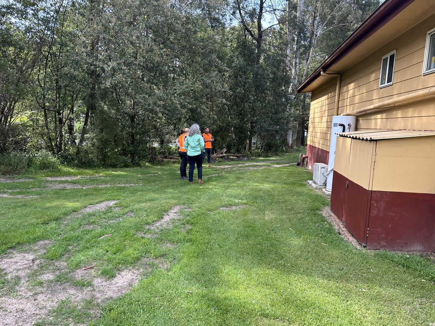 Three people from the site inspection standing on the lawn beside the Puffing Billy Railway building in Belgrave, with bushland behind and the downpipes and hot water unit along the building wall.